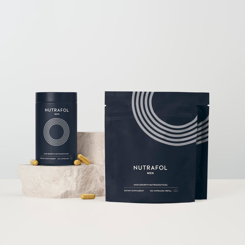 Nutrafol Men dietary supplement packaging, including a cylindrical tin and a stand-up pouch, both in dark navy blue with white graphics and text, displayed on a light surface with a small stone prop and scattered capsules.