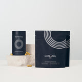 Nutrafol Men dietary supplement packaging, including a cylindrical tin and a stand-up pouch, both in dark navy blue with white graphics and text, displayed on a light surface with a small stone prop and scattered capsules.