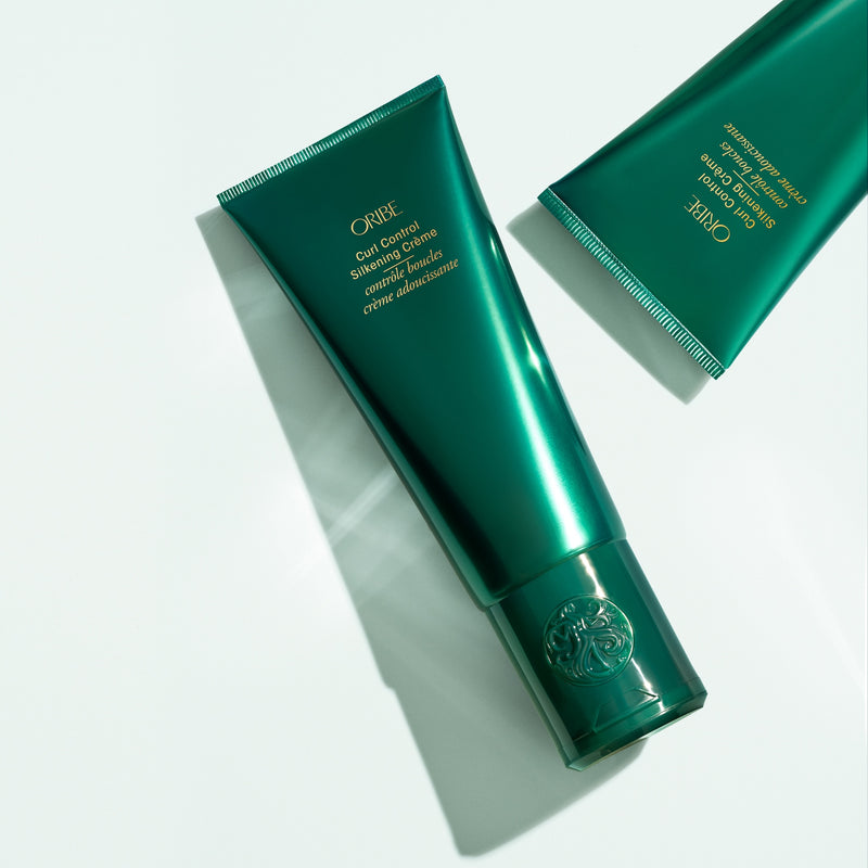 Two Oribe Curl Control Silkening Crème tubes arranged diagonally on a pale studio surface with crisp directional shadows