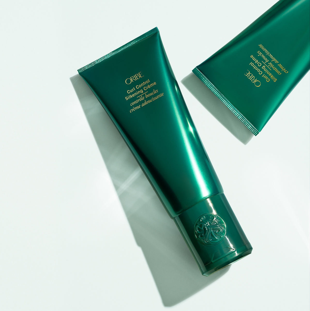 Two Oribe Curl Control Silkening Crème tubes arranged diagonally on a pale studio surface with crisp directional shadows