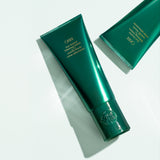 Two Oribe Curl Control Silkening Crème tubes arranged diagonally on a pale studio surface with crisp directional shadows
