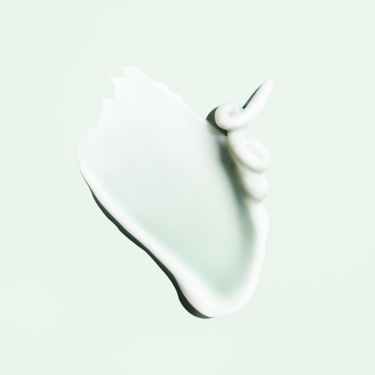 Glossy white cream texture of Oribe Curl Control Silkening Crème spread into a soft sweep with curled peaks on a pale green surface