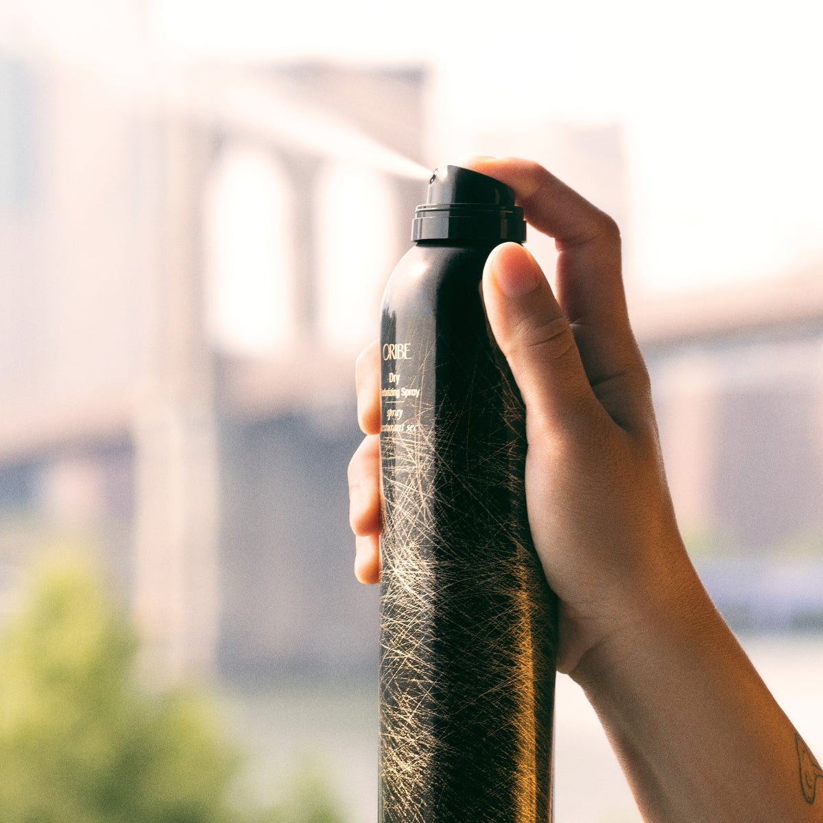 Hand pressing the exposed nozzle of an Oribe Dry Texturizing Spray 8.5 oz can in warm natural light