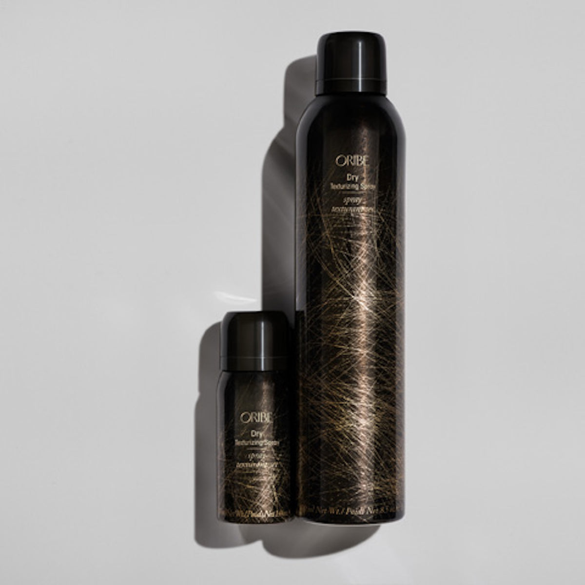 Oribe Dry Texturizing Spray standard and travel cans arranged closely on a gray studio surface with dramatic shadows and reflective gold linework