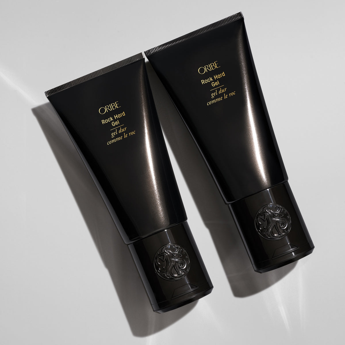 Two Oribe Rock Hard Gel black tubes arranged diagonally on a light gray studio surface with sharp directional shadows