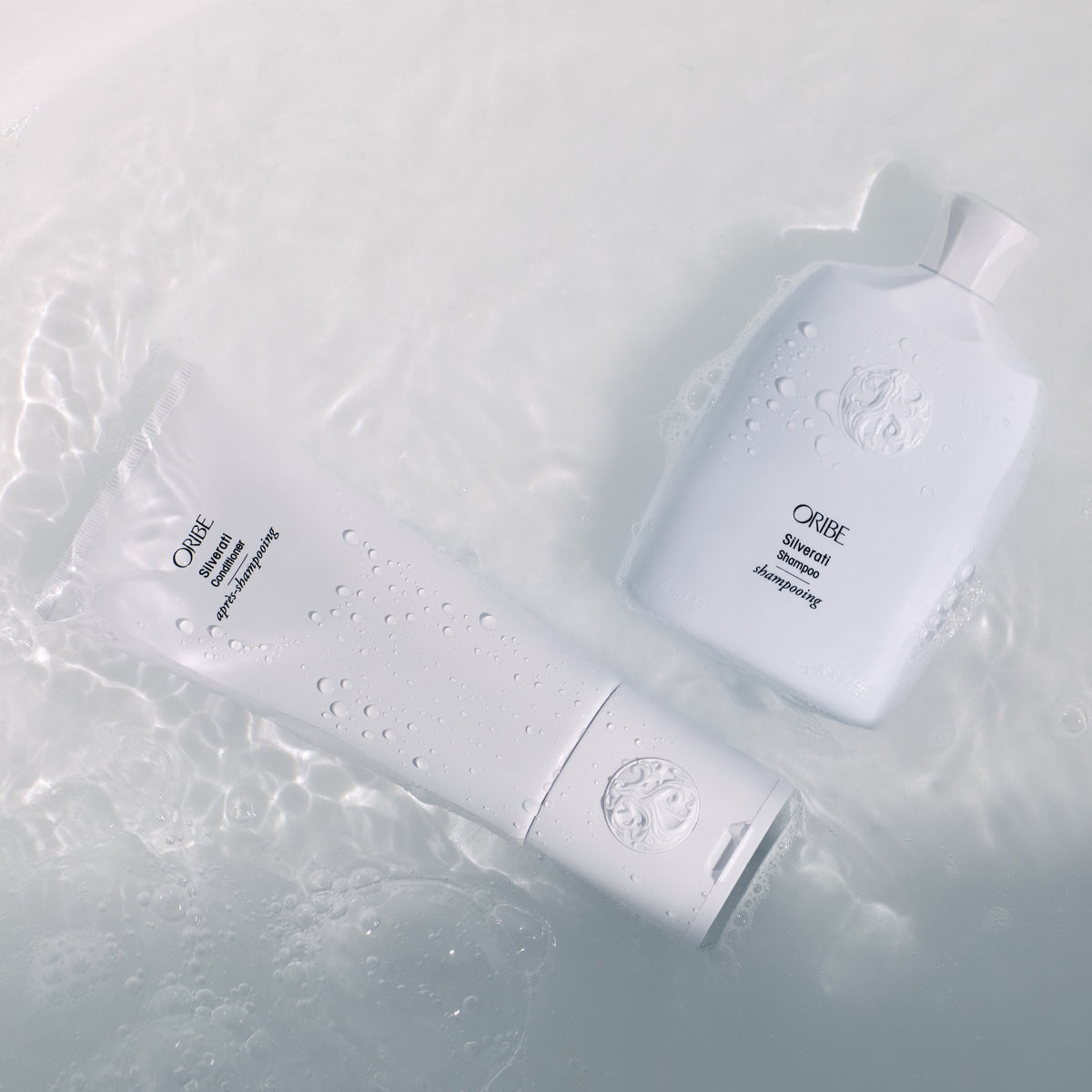 Oribe Silverati Shampoo and Conditioner resting in shallow water with droplets across the sculpted white packaging