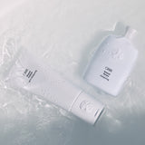 Oribe Silverati Shampoo and Conditioner resting in shallow water with droplets across the sculpted white packaging