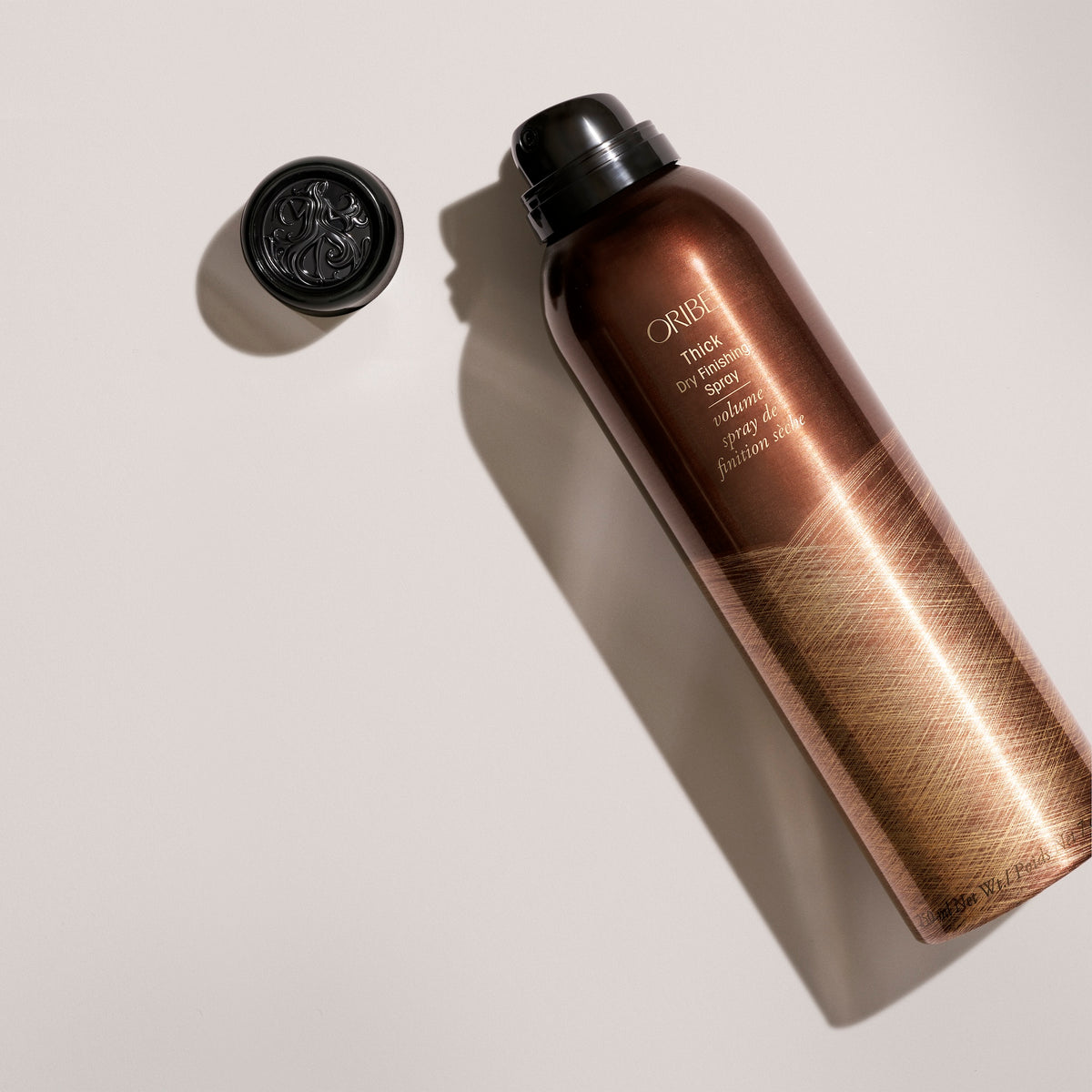 Oribe Thick Dry Finishing Spray standard 7 oz bronze can shown uncapped and angled on a pale gray studio surface with its embossed black cap and elongated directional shadow