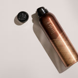 Oribe Thick Dry Finishing Spray standard 7 oz bronze can shown uncapped and angled on a pale gray studio surface with its embossed black cap and elongated directional shadow